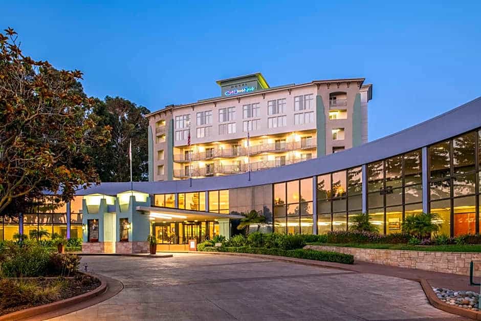 Modern hotel building with large windows and balconies, surrounded by landscaping, at dusk with lights illuminating the entrance.